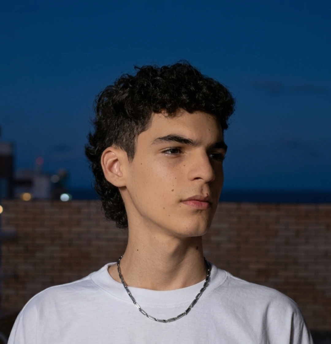 Retrato fotográfico de Kauã Cavalcante. Um jovem moreno com cabelo cacheado curto em corte mullet, usando uma camiseta branca básica e uma corrente de metal prata. Ele está posicionado em três quartos de perfil, olhando para a direita com expressão serena. O fundo é um cenário noturno urbano com céu azul escuro e um muro de tijolos ao fundo, com a iluminação natural da noite destacando seu rosto.